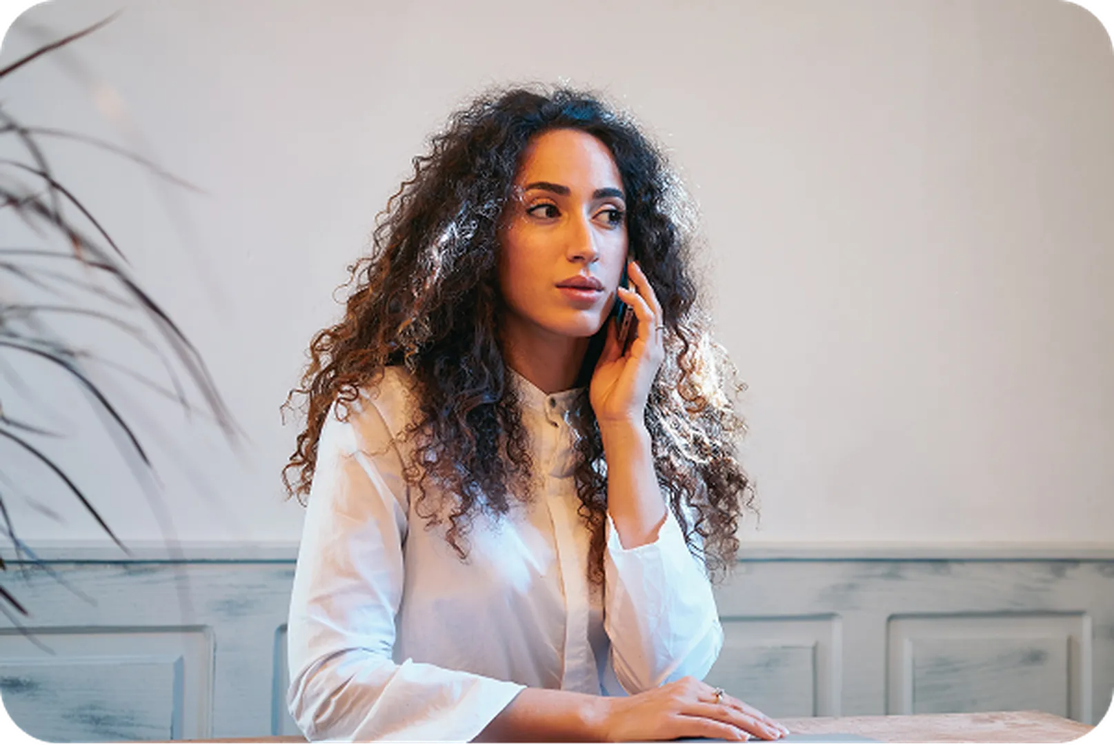 Woman with curly hair in white shirt, hand on face, seated at table with softly lit background