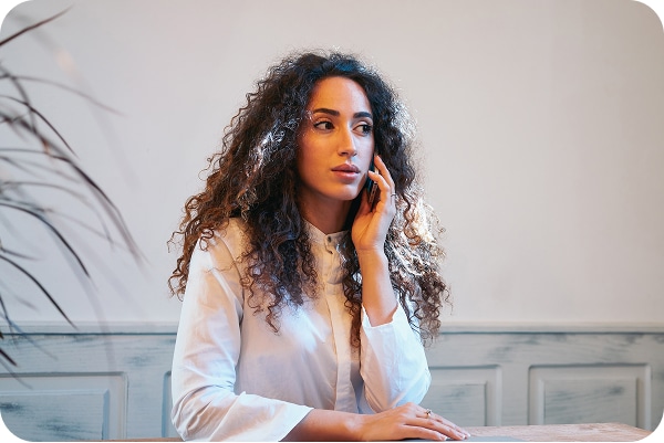 Woman with curly hair in white shirt, hand on face, seated at table with softly lit background