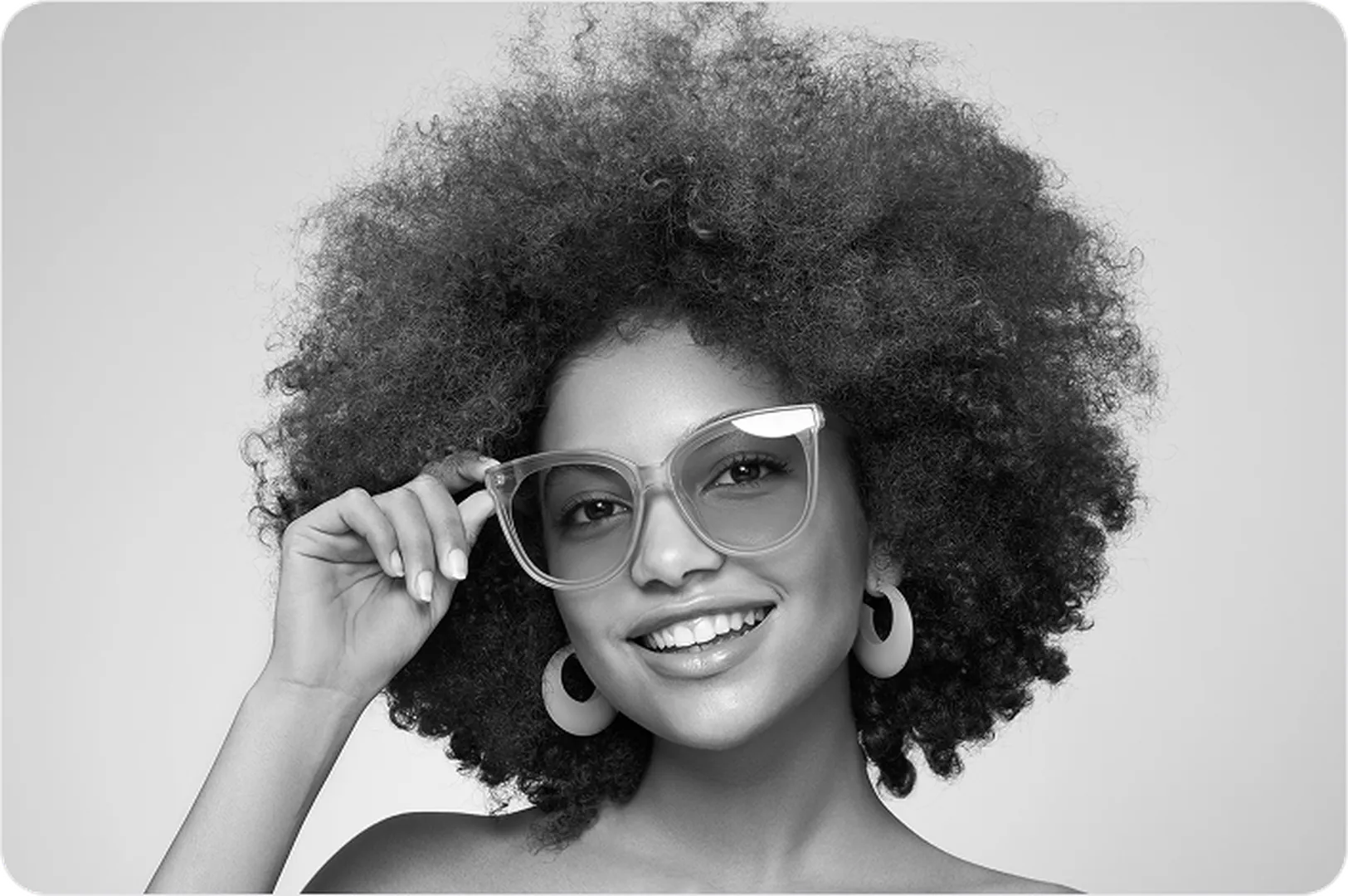 A woman with voluminous hair and glasses poses in front of a light silver backdrop.