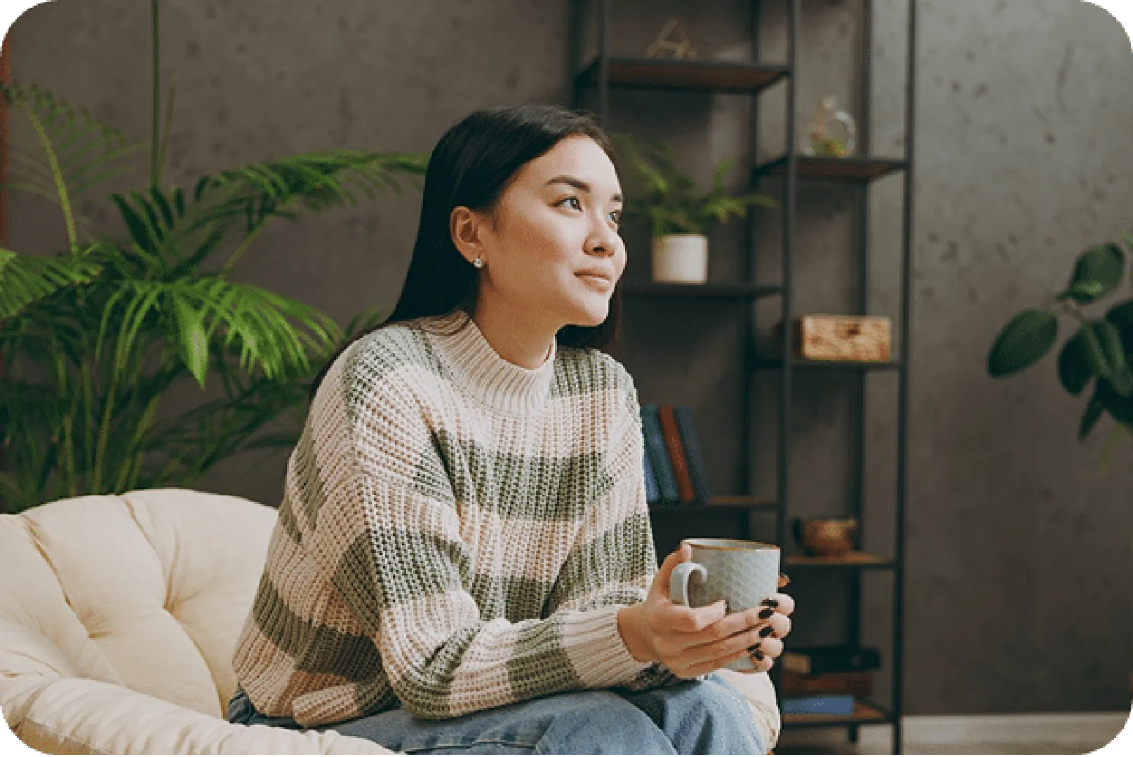 Person in cozy sweater holding a mug, seated near indoor plants and modern bookshelf.