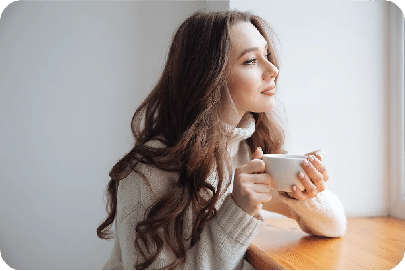 Person with long wavy hair by window, holding white mug, wearing cozy sweater, sunlight streaming.
