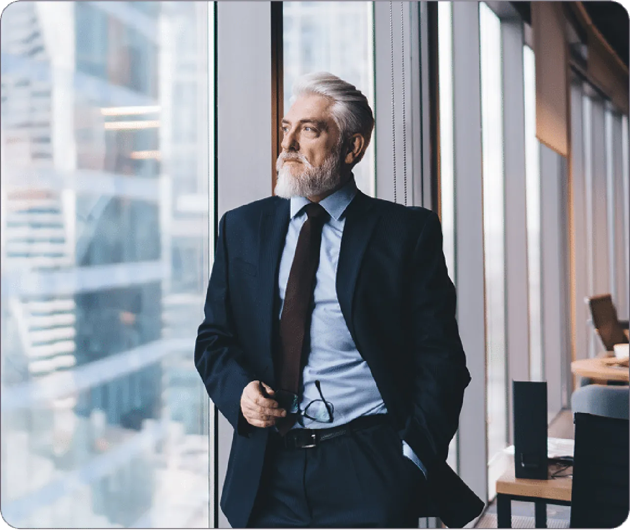 Man in dark suit stands by tall window, holding glasses, overlooking city skyline thoughtfully.