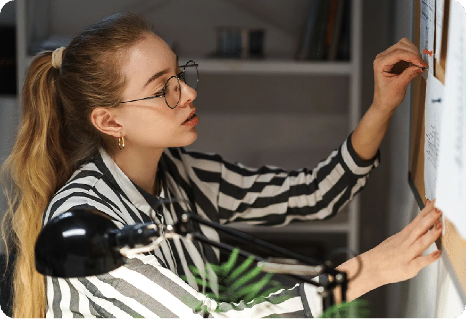 Person in striped shirt pins notes on bulletin board under desk lamp in a cozy setting.